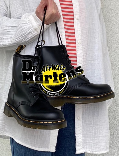 Frau mit Jeans weißem Hemd und geringeltem T-Shirt hält an den Schnürsenkeln einen Dr. Martens Schuh in Schwarz. Auf dem Bild ist auch das Dr. Martens Logo dargestellt.