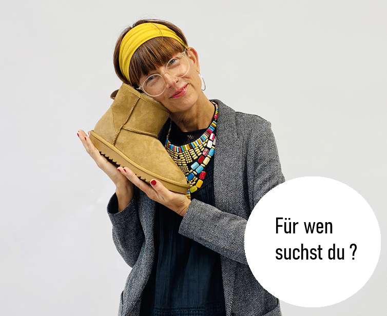 Junge Frau mit Jacke Jeanshemd und geringeltem T-Shirt steht mit einem hohen Converse mit den Beinen verschänkt an einer Wand, Text Für wen suchst du ?