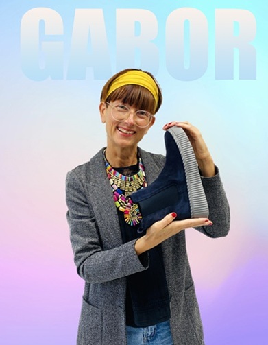 Frau in schwarzer Kleidung und blonden Haaren hat einen Gabor Sneaker in der Hand und zeigt auf diesen. Es ist ein Gabor Sneaker in weiß mit Straßteinen vorne, Bild hat Verlinkung zu dem Schuh im Onlineshop
