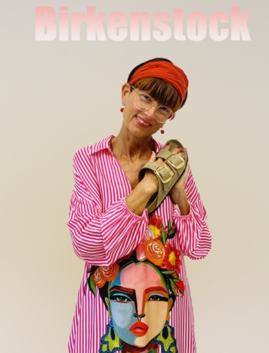 Frau mit blauem Pullover und Jeans hat die Hand in einem Paar Birkenstock Pantoletten in grün und brauner Farbe. Dargestellt in einem Bild mit weißem Hintergund. Das Bild steht auf dem Boden. Auf dem Bild wird auch ein Birkenstock Logo dargestellt und man gelangt per Link auf alle Birkenstock Schuhe