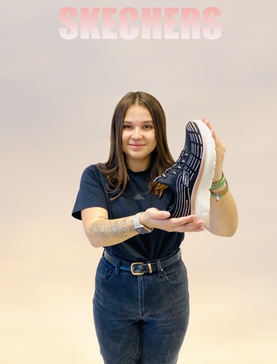 Frau in schwarzer Kleidung und blonden Haaren hat einen Gabor Sneaker in der Hand und zeigt auf diesen. Es ist ein Gabor Sneaker in weiß mit Straßteinen vorne, Bild hat Verlinkung zu dem Schuh im Onlineshop