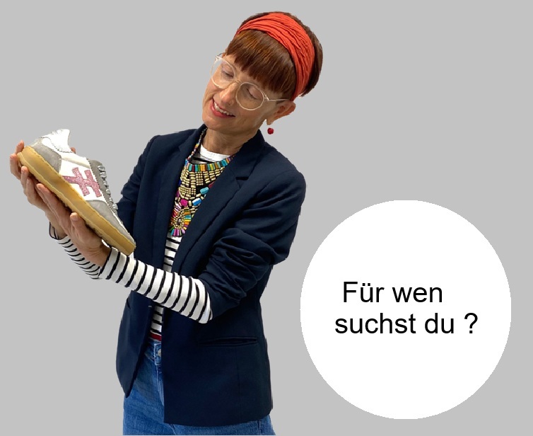 Frau mit blauem Blazer geringeltem Shirt bunter Kette und orangefarbenen Stirnband und Jean hält einen Another Trend Sneaker in der Hand und bewundert ihn neben Ir ist ein Kreis mit text Für wen suchst du neu