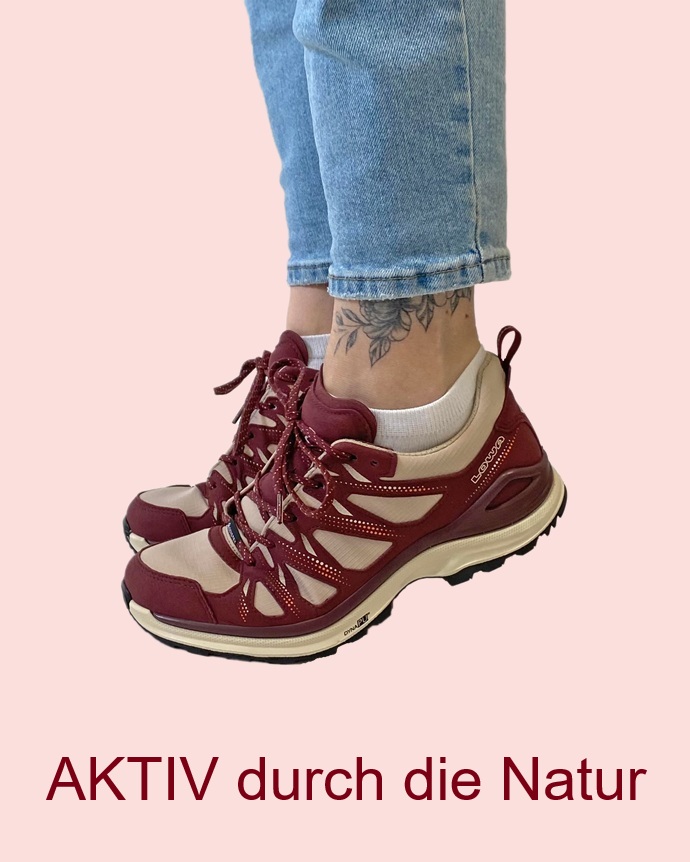 Frau Springt mit Ihrem Leichtwanderschuh nach oben der Schuh hat die Farben Bordeaux und beige und heller Sohle duie Frau hat eine Jeans an und weiße Socken und ein Tatoo